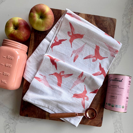 White towel with pink bird pattern on a wooden cutting board with apples and a pink container.
