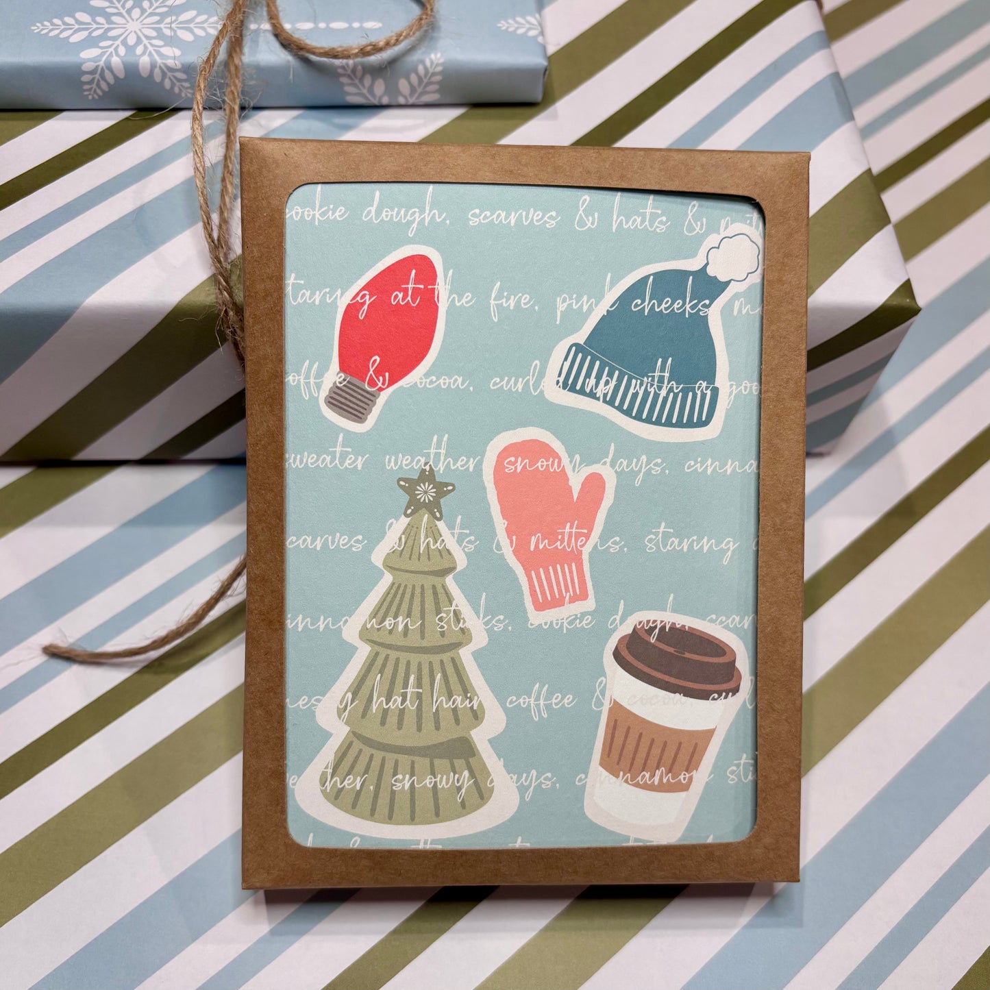 Box of Christmas-themed cards with illustrations of a red light bulb, green tree, pink mittens, and coffee cup on a striped background.