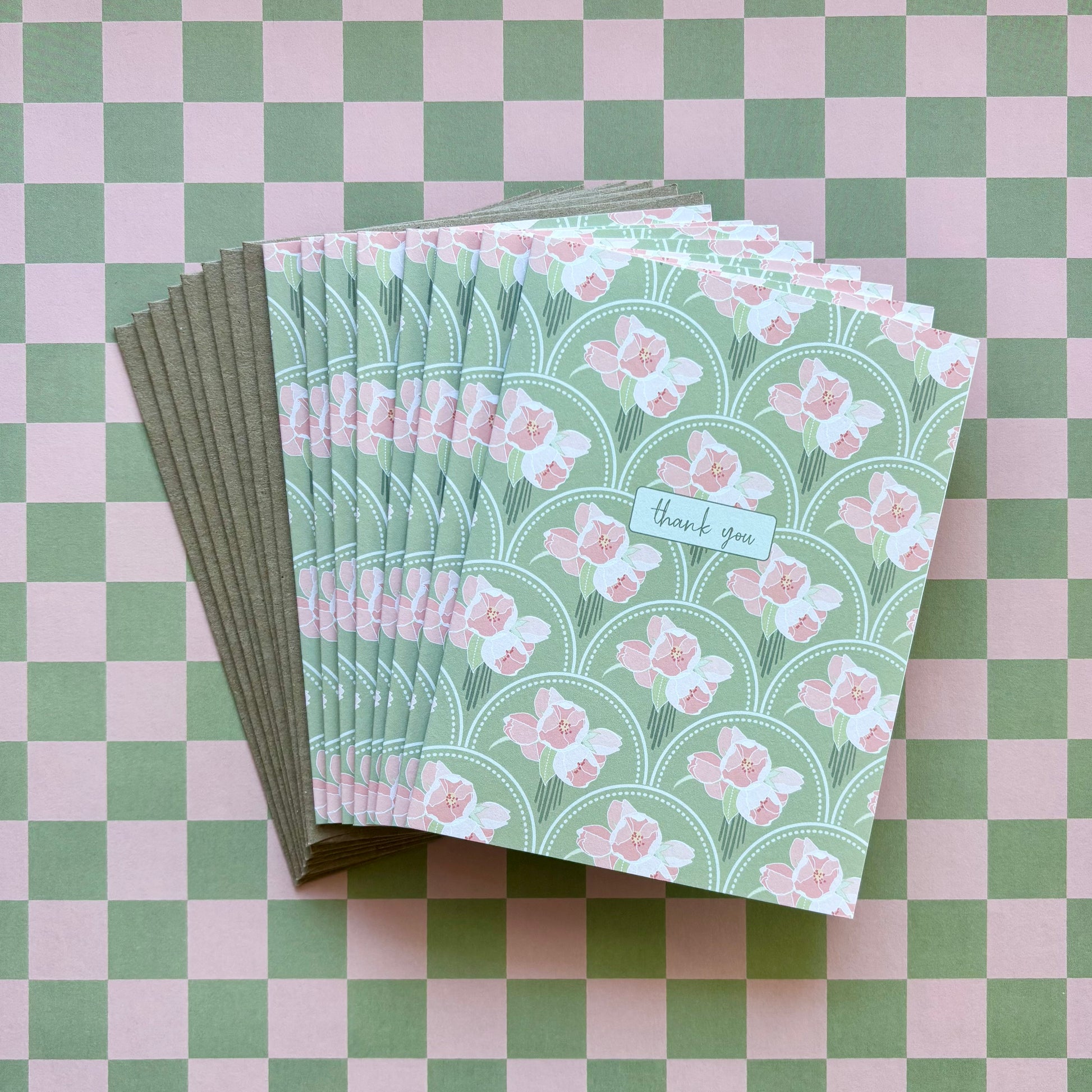 Fanned stack of eight thank you greeting cards with pink flowers on a green scallop pattern background and kraft brown envelopes