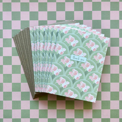 Fanned stack of eight thank you greeting cards with pink flowers on a green scallop pattern background and kraft brown envelopes