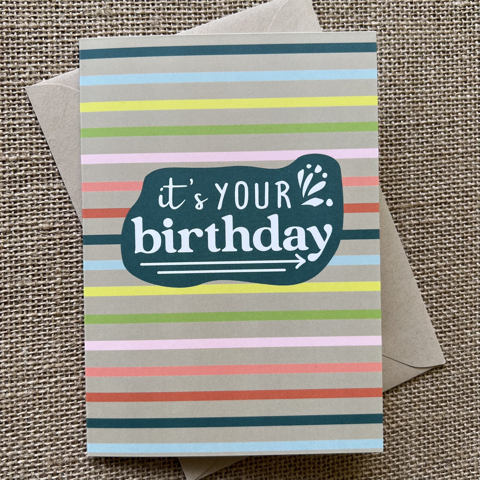 Rainbow striped background with a teal organic shape, saying "It's your birthday" in white type, show with kraft paper envelope