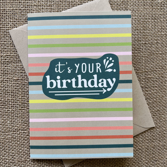 Rainbow striped background with a teal organic shape, saying "It's your birthday" in white type, show with kraft paper envelope