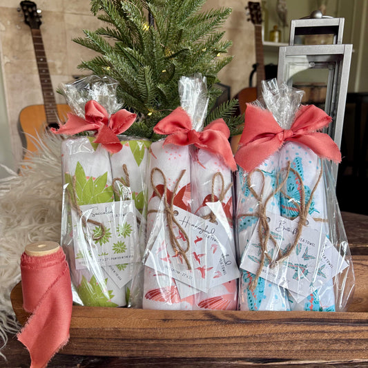 Three clear bags each containing two tea towels and tied with an orange silk ribbon bow, in front of a mini Christmas tree