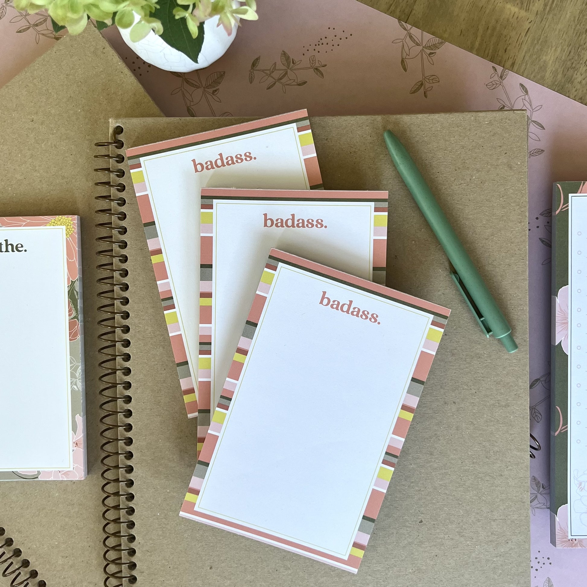 Stack of three badass notepads, styled with a green pen and other stationery items