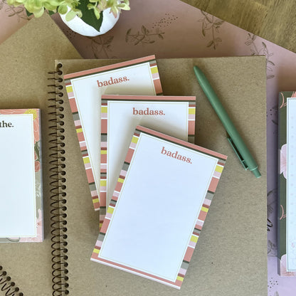 Stack of three badass notepads, styled with a green pen and other stationery items