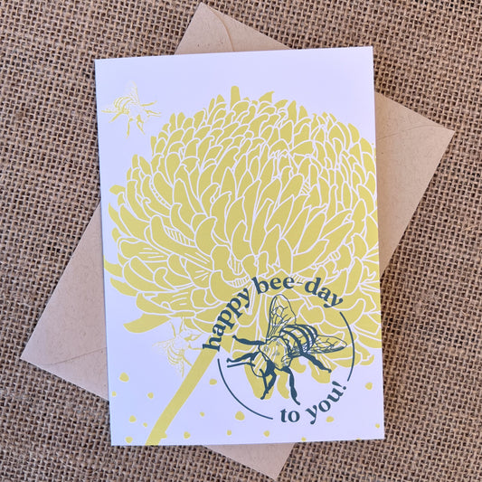 Greeting card with white background, citron chrysanthemum flower and teal bee illustration and text reading "happy bee-day to you", styled with a kraft envelope on a tan burlap background