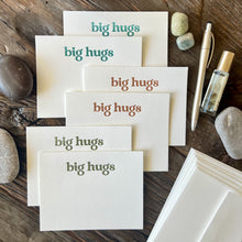 Load image into Gallery viewer, Staggered pile of six flat notecards, white in color with the words &quot;big hugs&quot; letterpress printed with embossed texture, two cards in teal ink, two in a warm orange and two in olive green. Styled on a wooden table with river rocks, a gold pen and a bottle of essential oil.
