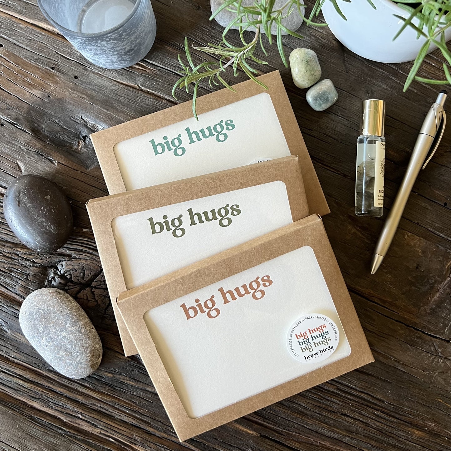 Three craft paper boxes with clear windows showing printed notecards with the words "big hugs" in teal, olive and warm orange ink. Styled on a wooden table with river rocks, a gold pen, a bottle of essential oils and a small peak of rosemary branches. 