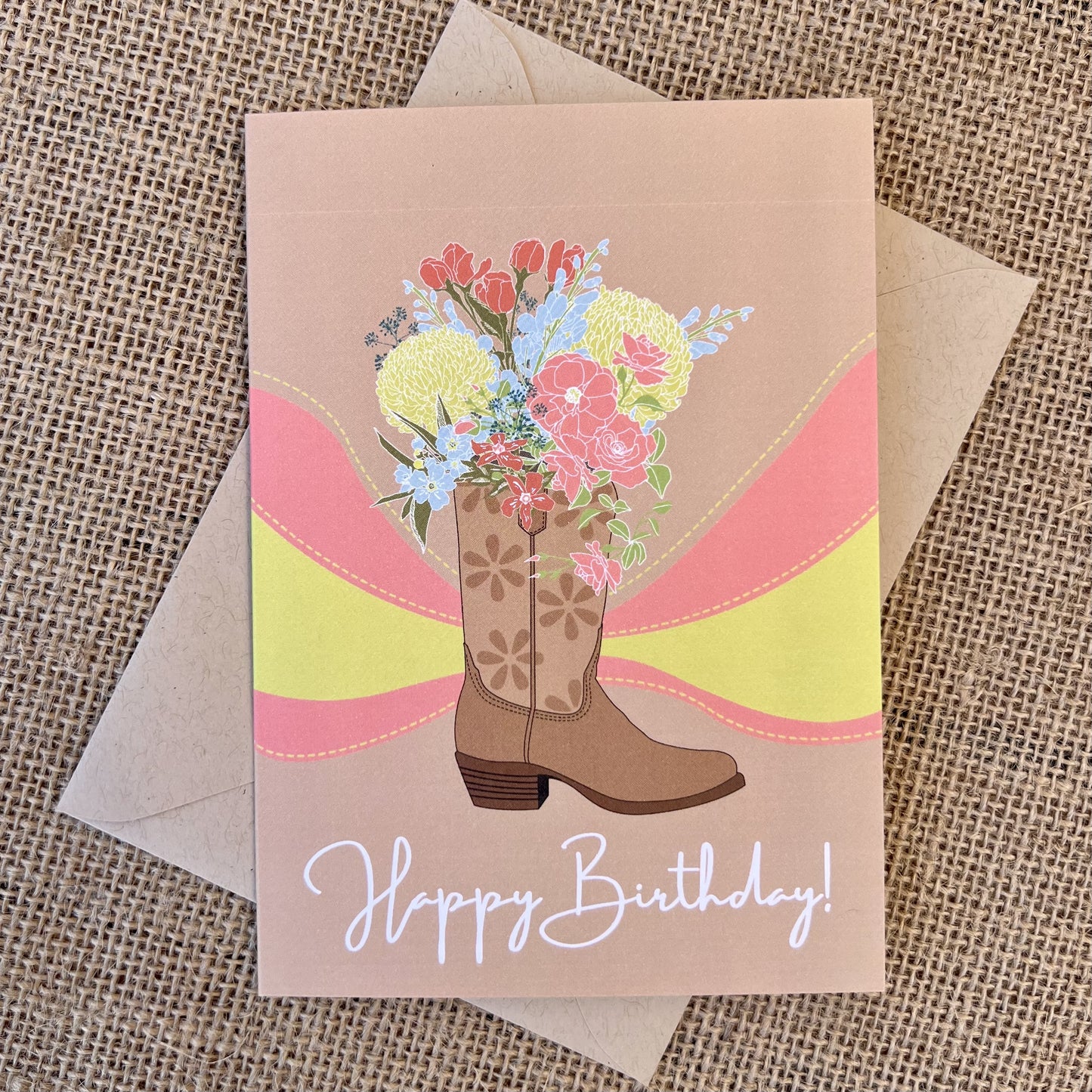 Greeting card with tan background featuring a cowboy boot illustration with colorful flowers, text reading "happy birthday", styled with a kraft envelope on a tan burlap background