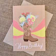 Load image into Gallery viewer, Greeting card with tan background featuring a cowboy boot illustration with colorful flowers, text reading &quot;happy birthday&quot;, styled with a kraft envelope on a tan burlap background
