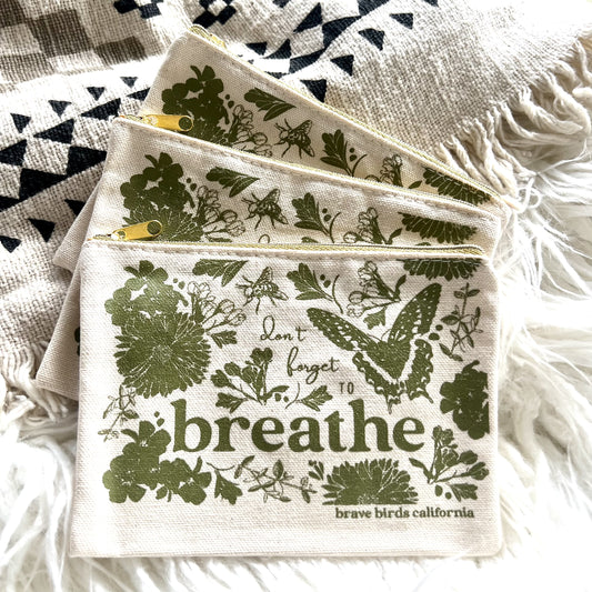 Stack of three zip pouches in natural canvas with olive green print and gold zippers. Print reads "don't forget to breathe"