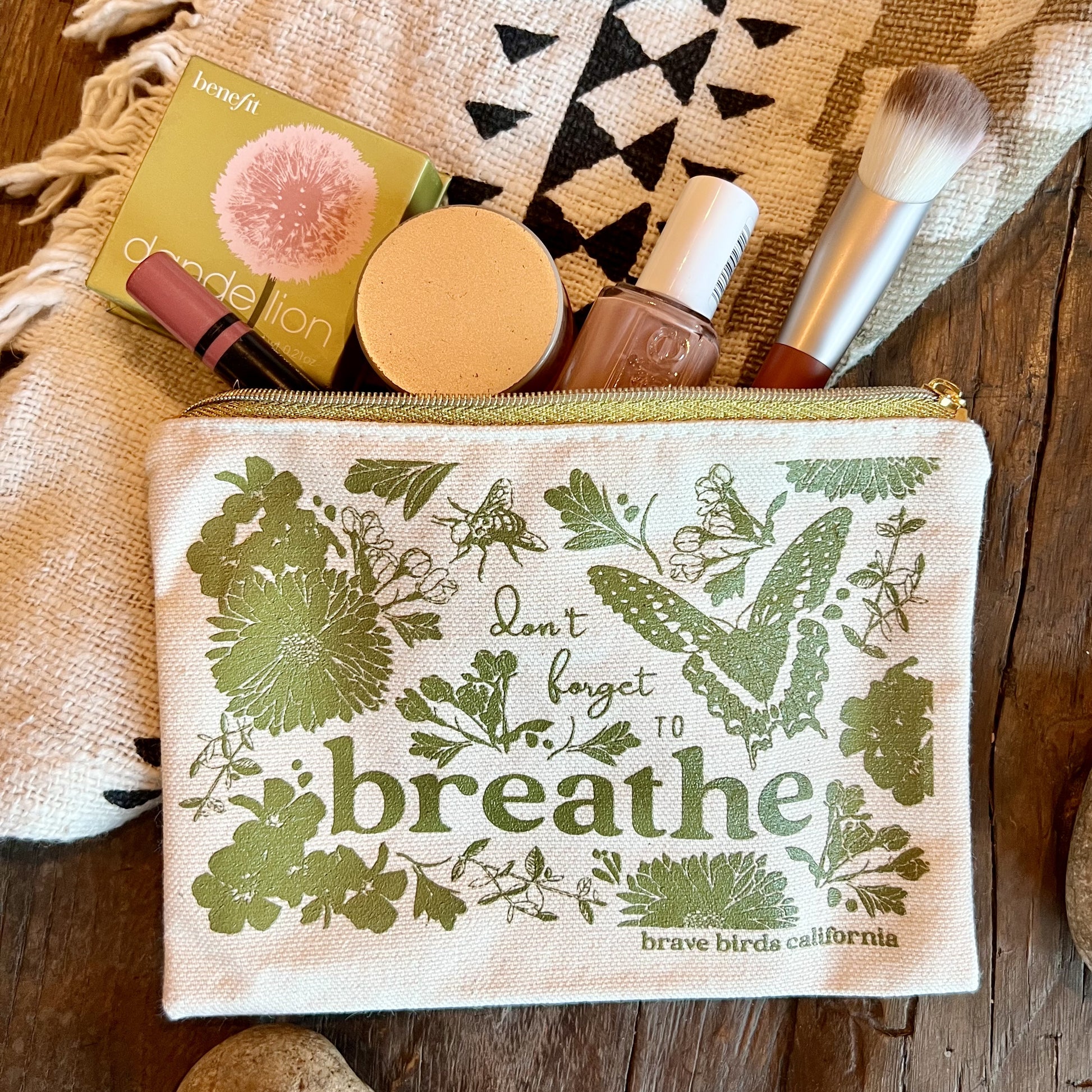 Zip pouch in natural canvas with olive green print and gold zippers, shown open with makeup items peeking out. Print reads "don't forget to breathe"