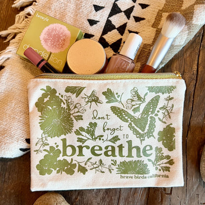 Zip pouch in natural canvas with olive green print and gold zippers, shown open with makeup items peeking out. Print reads "don't forget to breathe"
