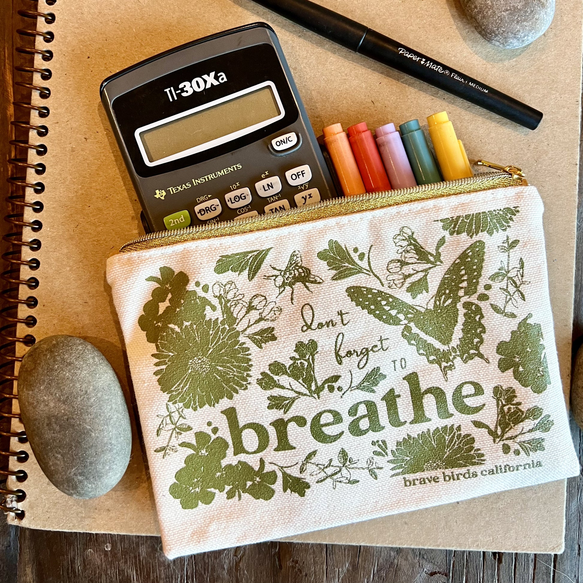 Zip pouch in natural canvas with olive green print and gold zippers, shown open with a calculator and markers peeking out. Print reads "don't forget to breathe"