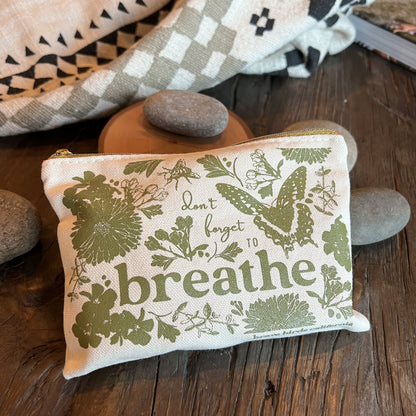 Zip pouch in natural canvas with olive green print and gold zippers, shown zipped closed with items inside. Print reads "don't forget to breathe"