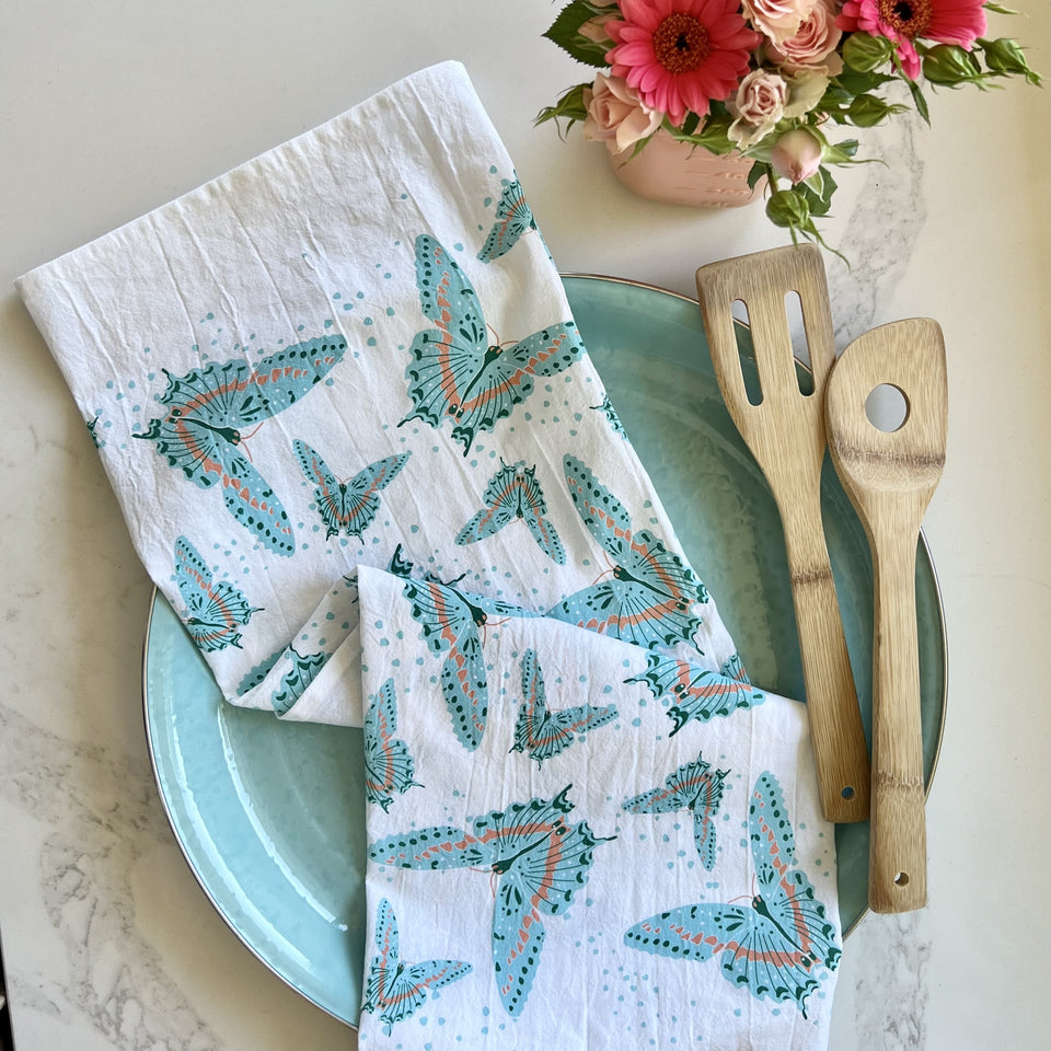 White cotton tea towel printed with a blue butterfly pattern, styled on a blue plate with wooden cooking utensils