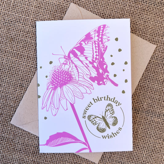 Greeting card with white background, dark pink butterfly and echinacea flower, and olive green text that reads "sweet birthday wishes", styled with a kraft envelope on tan burlap
