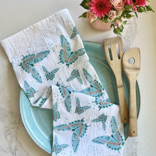 White cotton tea towel printed with a blue butterfly pattern, styled on a blue plate with wooden cooking utensils