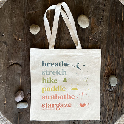 Natural Canvas Tote with rainbow colored ink and the words breathe, stretch, hike, paddle, sunbathe, stargaze, each in a different color. Shown flat on a rustic wood surface with river rocks scattered around.