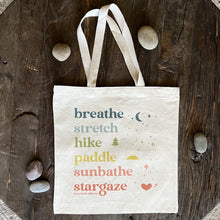 Load image into Gallery viewer, Natural Canvas Tote with rainbow colored ink and the words breathe, stretch, hike, paddle, sunbathe, stargaze, each in a different color. Shown flat on a rustic wood surface with river rocks scattered around.
