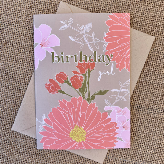 Greeting card with khaki background and pink flowers, text reading "birthday girl", styled with a kraft envelope on a tan burlap background