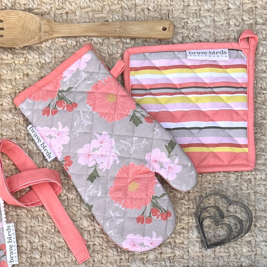 Floral printed oven mitt in pink & khaki with a color-coordinated striped square potholder, both with hanging loops, styled with a wooden spoon and heart shaped cookie cutters