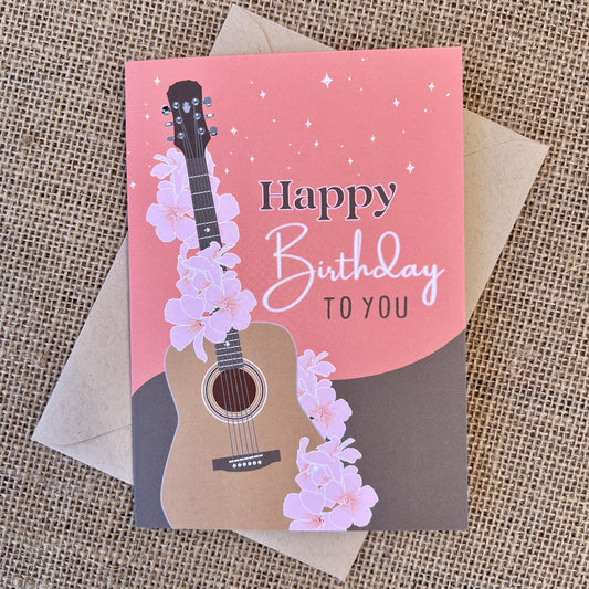 Greeting card with pink & brown background and pink floral guitar illustration, text reading "happy birthday to you", styled with a kraft envelope on a tan burlap background