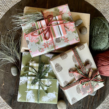 Load image into Gallery viewer, Pile of three giftwrapped boxes in pine branch and pinecone patterns, in pink, olive and brown color palette and tied with yarn
