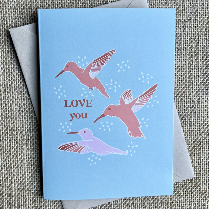 Greeting card with a light blue background, three pink hummingbirds, and the text reading 