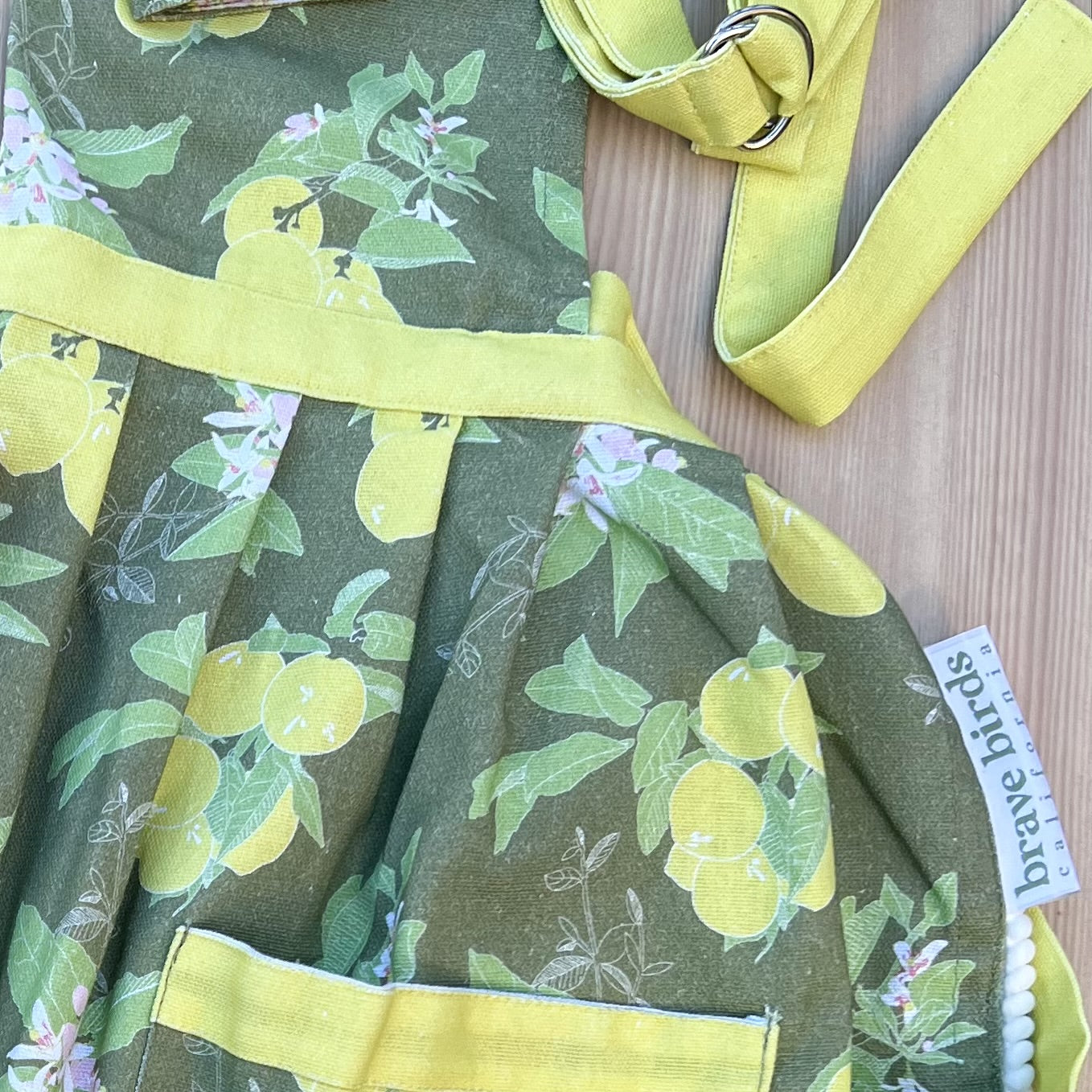 Zoomed in detail shot of apron trim: pleating at the waist, citron trim, woven label reading "brave birds california" and white scalloped fabric edging