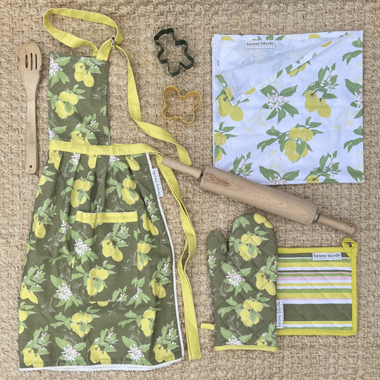 Collection of lemon print kitchen textiles in olive green, citron yellow and white color palette. Apron, oven mitt, potholder and tea towel, styled with a rolling pink, wooden spoon and cookie cutters.