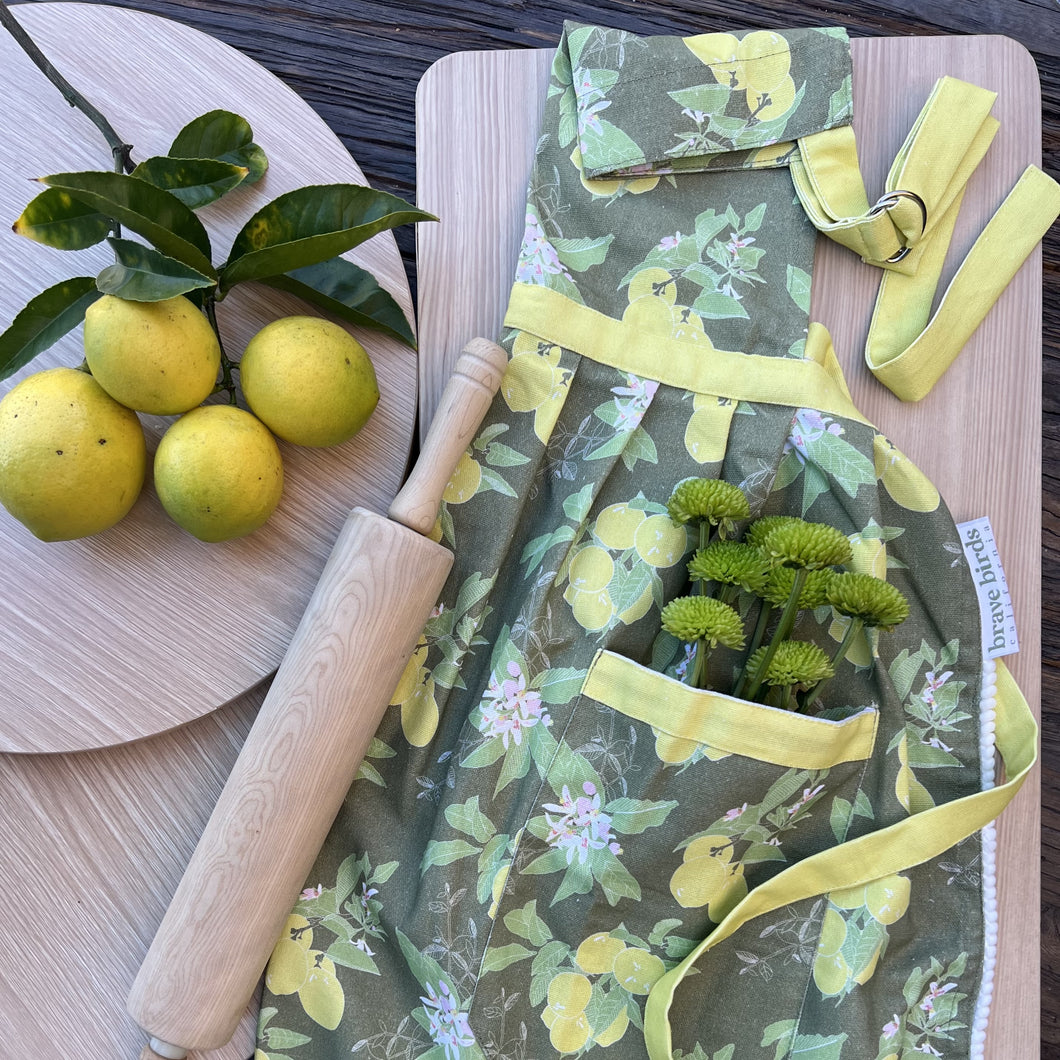 Cotton apron with a lemon print in olive green and citron yellow, yellow tie accents, styled with a rolling pin, four lemons and green flowers in the pocket