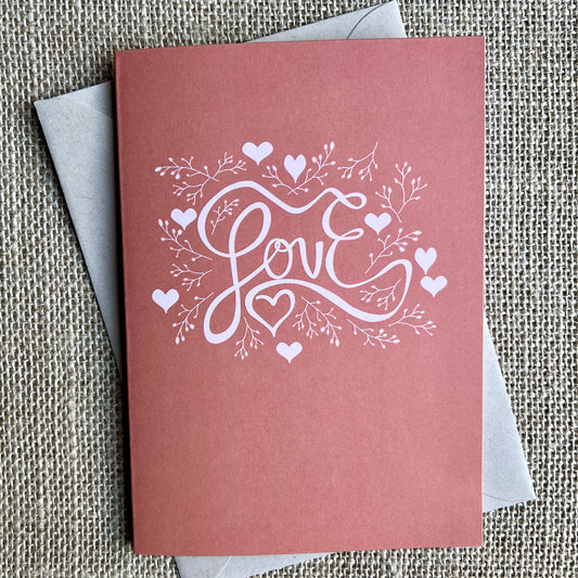 Greeting card with a warm red/pink background and a hand illustrated "Love" with hearts and branches in light pink