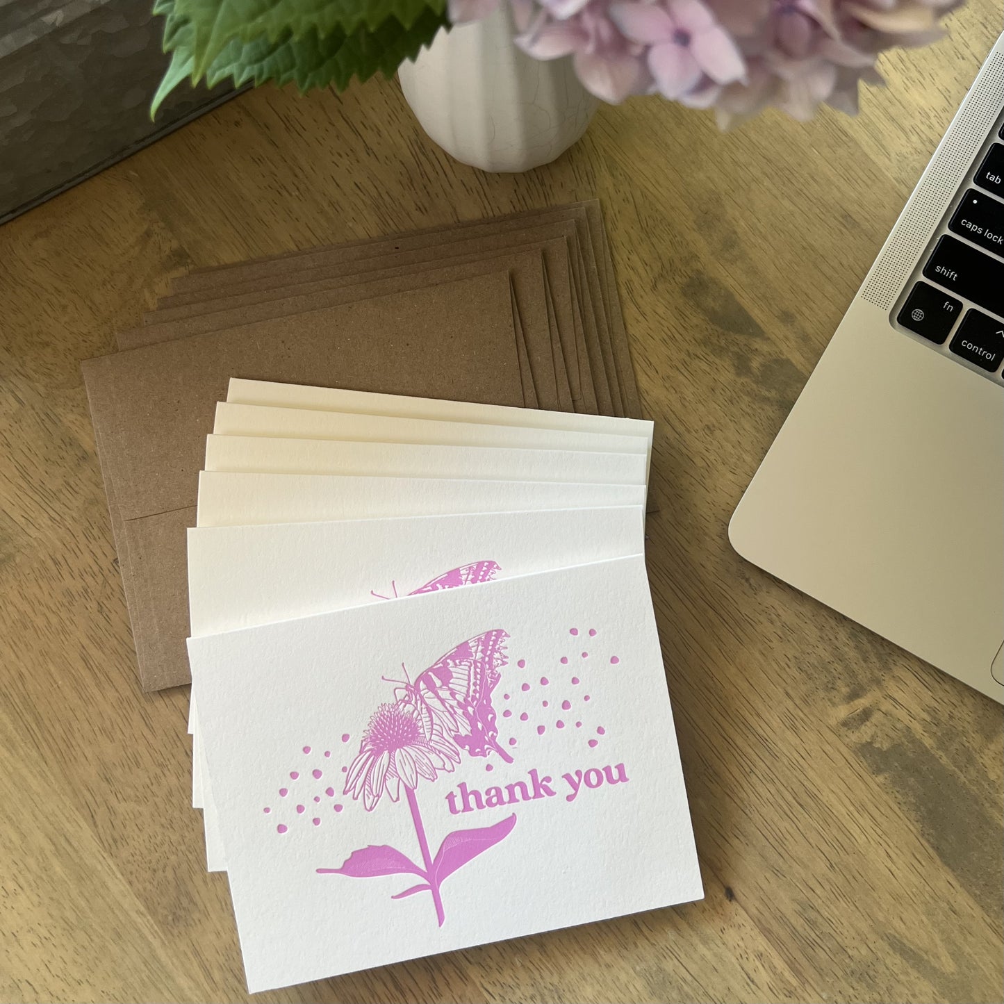 Stack of pink butterfly letterpress thank you cards with kraft envelopes, styled on a wooden desk with a laptop computer and pink hydrangea flowers
