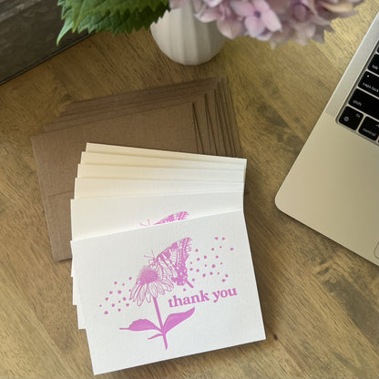 Stack of pink butterfly letterpress thank you cards with kraft envelopes, styled on a wooden desk with a laptop computer and pink hydrangea flowers