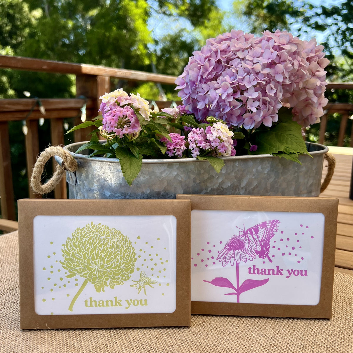 Stack of kraft paper boxes with windows showing two thank you note artworks - one print of a butterfly and echinacea flower in dark pink and one showing a chrysanthemum and a bee in citron yellow, styled in front of a bouquet of hydrangeas