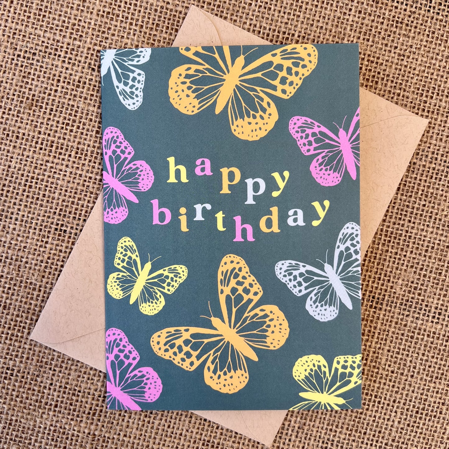 Greeting card with teal background and pink, orange and blue butterflies, text reading "happy birthday", styled with a kraft envelope on a tan burlap background