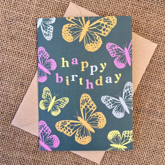 Greeting card with teal background and pink, orange and blue butterflies, text reading "happy birthday", styled with a kraft envelope on a tan burlap background