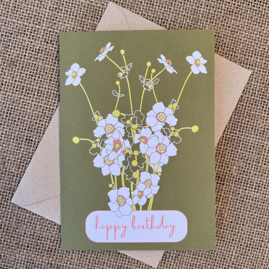 Greeting card with olive green background and pink/white flowers, text reading "happy birthday", styled with a kraft envelope on a tan burlap background
