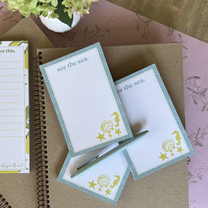 Stack of three see the sea notepads, styled with a blue pen and other stationery items