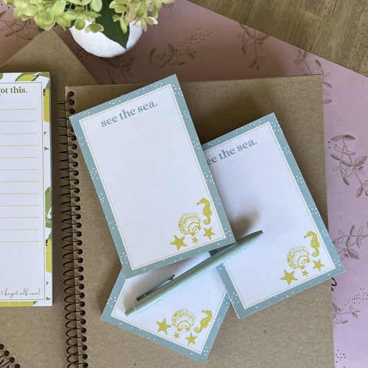Stack of three see the sea notepads, styled with a blue pen and other stationery items