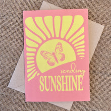 Load image into Gallery viewer, Greeting card with pink background and yellow sunshine and butterfly illustration, text reading &quot;sending sunshine&quot;, styled with a kraft envelope on a tan burlap background
