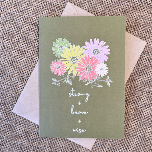 Greeting card with olive green background and colorful daisy flowers, text reading "strong + brave + wise", styled with a kraft envelope on a tan burlap background
