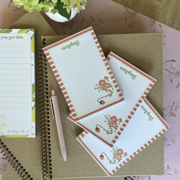 Stack of three unplug notepads, styled with a pink pen and other stationery items