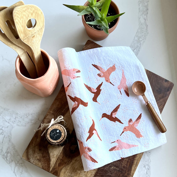 White tea towel with shades of rust & peach colored hummingbirds, styled with a wooden cutting board, a copper teaspoon, wooden spoons, a jar of fig jam, and a plant