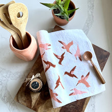 Load image into Gallery viewer, White tea towel with shades of rust &amp; peach colored hummingbirds, styled with a wooden cutting board, a copper teaspoon, wooden spoons, a jar of fig jam, and a plant
