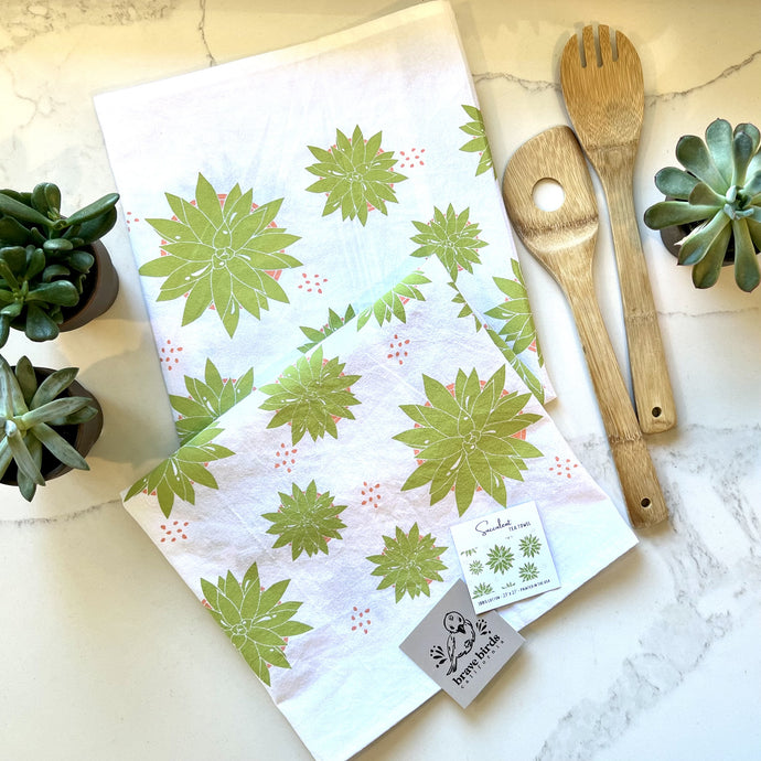 Succulent print tea towel folded on a kitchen counter, styled with wooden spoons and real succulent plants
